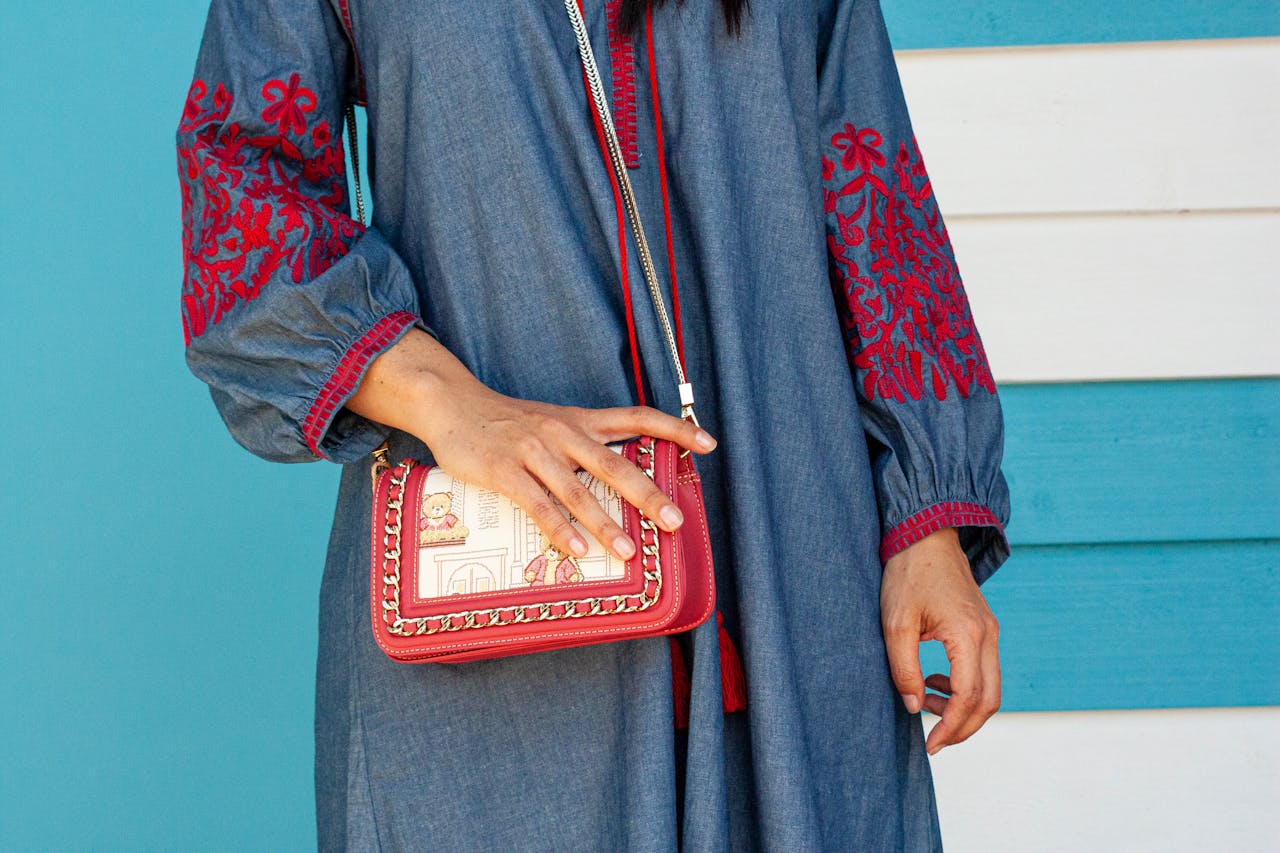 Close-up of a woman's hand on a red handbag, wearing a blue embroidered dress against a blue background.